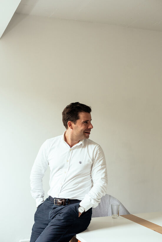 Felix Haas with white shirt and blue trousers sits at the edge of the table and looks to the side