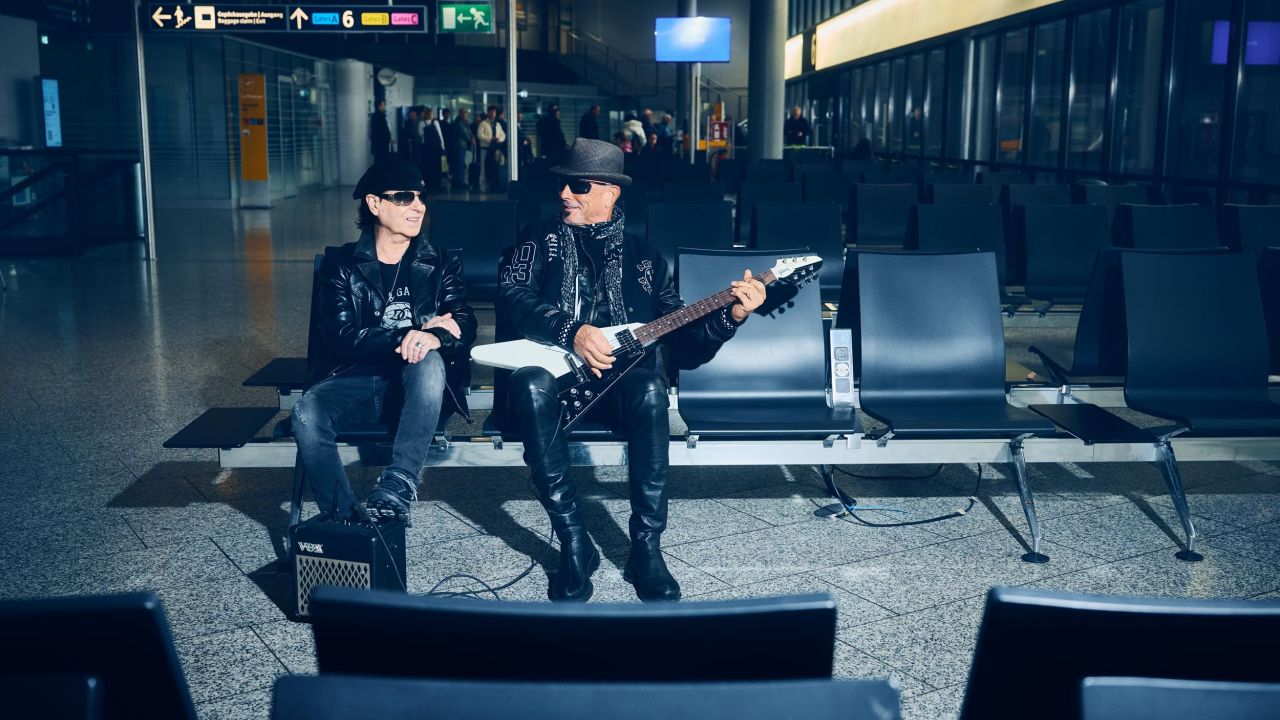 Zwei Bandmitglieder der Scorpions sitzen in einem Wartebereich am Flughafen, einer hält eine E-Gitarre
