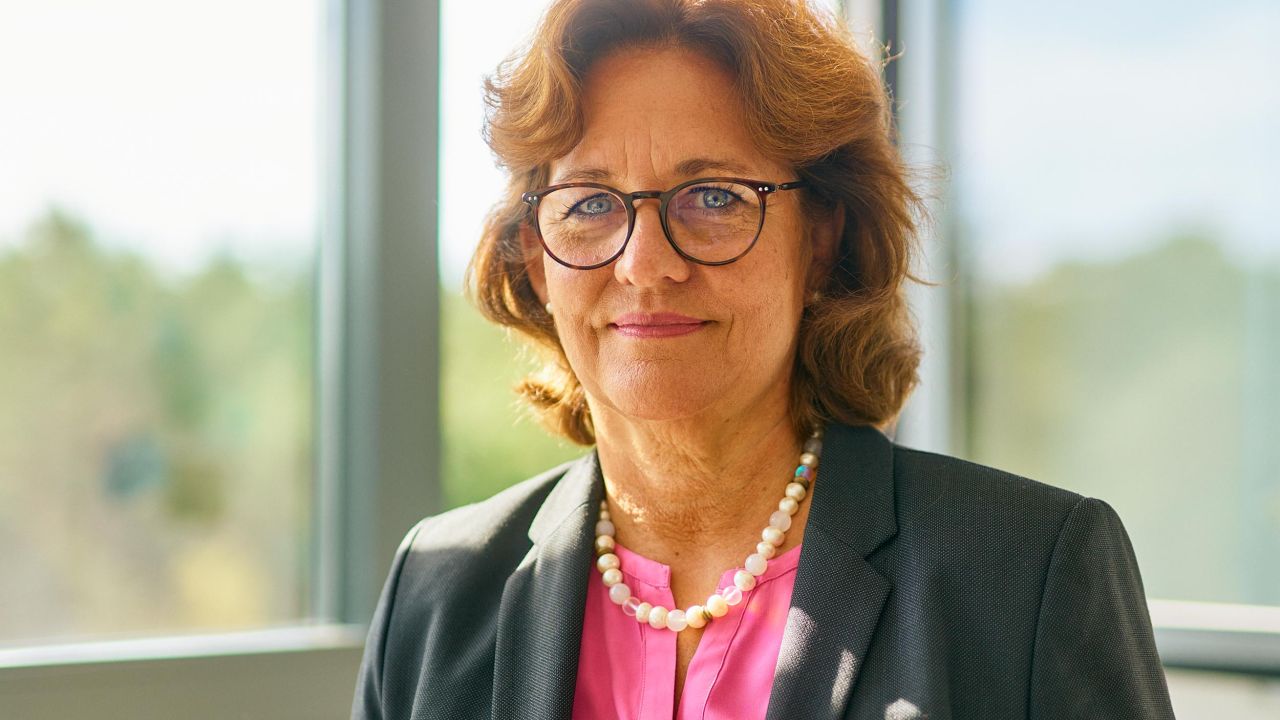 Nahaufnahme einer lächelnden Frau in Businesskleidung mit Brille und Perlenkette vor einem großen Fenster