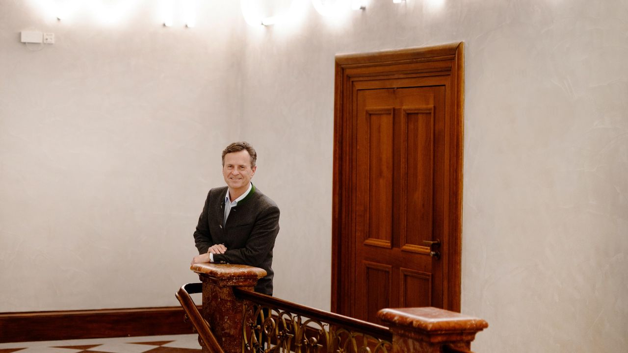 Ein lächelnder Mann steht am Treppengeländer in einem Foyer, im Hintergrund leuchten Neonbuchstaben das Wort „Wirtschaft“.