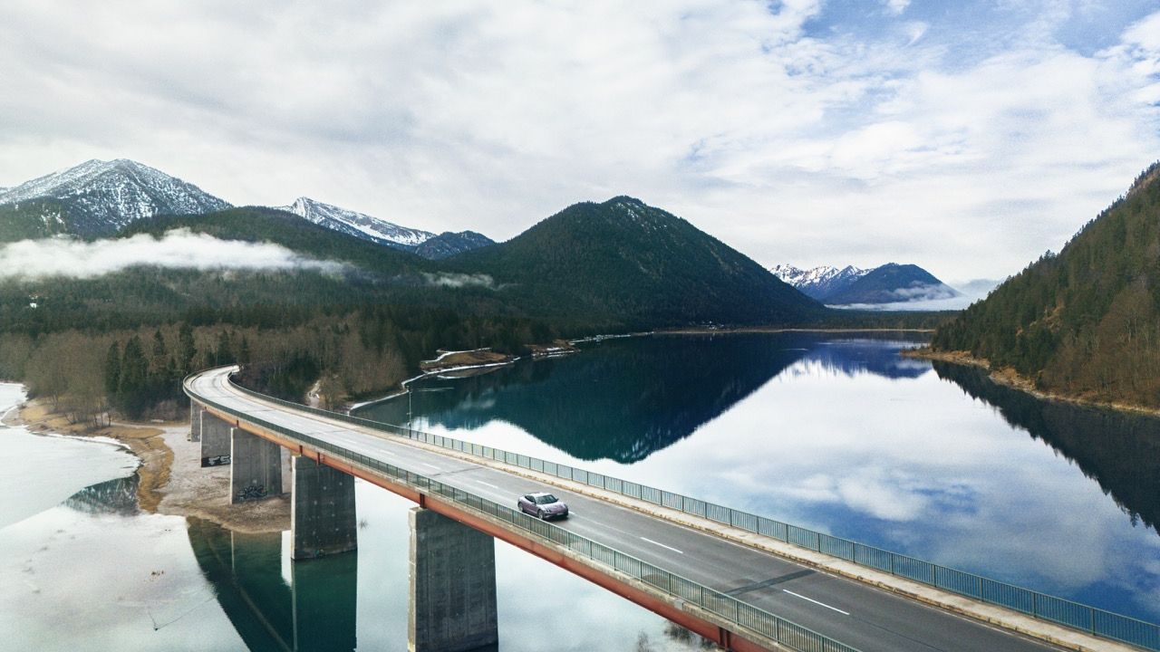 Aerial view of a Porsche Taycan 4 crossing a long bridge over an Alpine lake