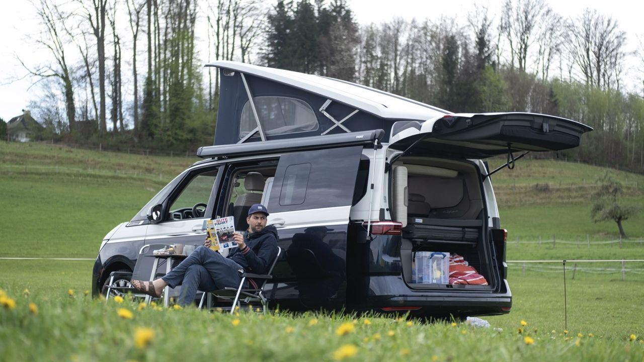 A man relaxes in a camping chair in front of his VW California with the pop-up roof and awning extended.