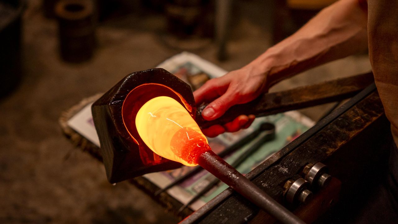 Close-up of a glassblower's hand shaping glowing hot, orange glass on a blowpipe using a tool in a dimly lit workshop Close-up of a glassblower's hand shaping glowing hot, orange glass on a blowpipe using a tool in a dimly lit workshop