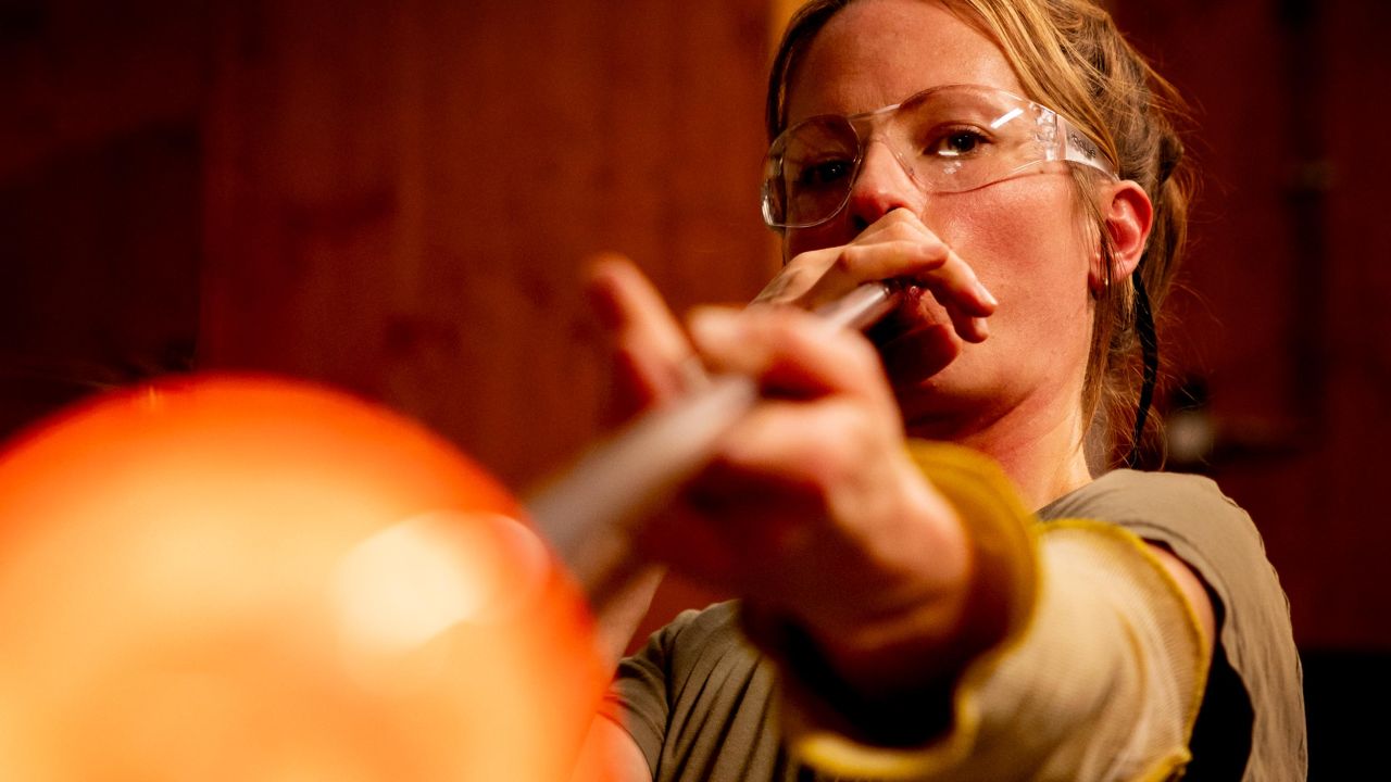 A female glassblower wearing safety goggles is blowing through a long blowpipe, the tip of which glows orange and is blurred in the foreground A female glassblower wearing safety goggles is blowing through a long blowpipe, the tip of which glows orange and is blurred in the foreground