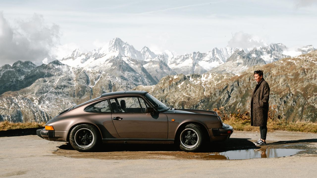 A brown Porsche 911 stands on a mountain road in front of a panorama of snow-covered Alpine peaks, with a woman standing next to the car. A brown Porsche 911 stands on a mountain road in front of a panorama of snow-covered Alpine peaks, with a woman standing next to the car.