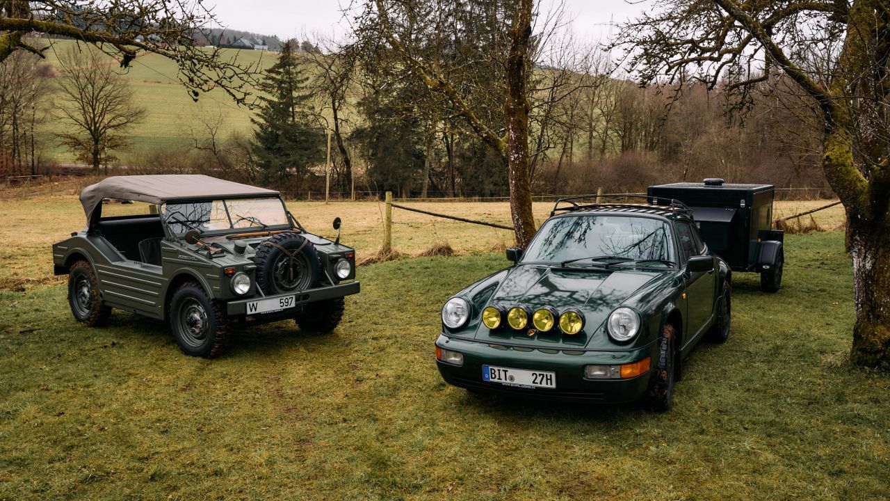 A dark green Porsche 911 with auxiliary headlights and a trailer is parked next to an olive green military off-road vehicle on a meadow. A dark green Porsche 911 with auxiliary headlights and a trailer is parked next to an olive green military off-road vehicle on a meadow.