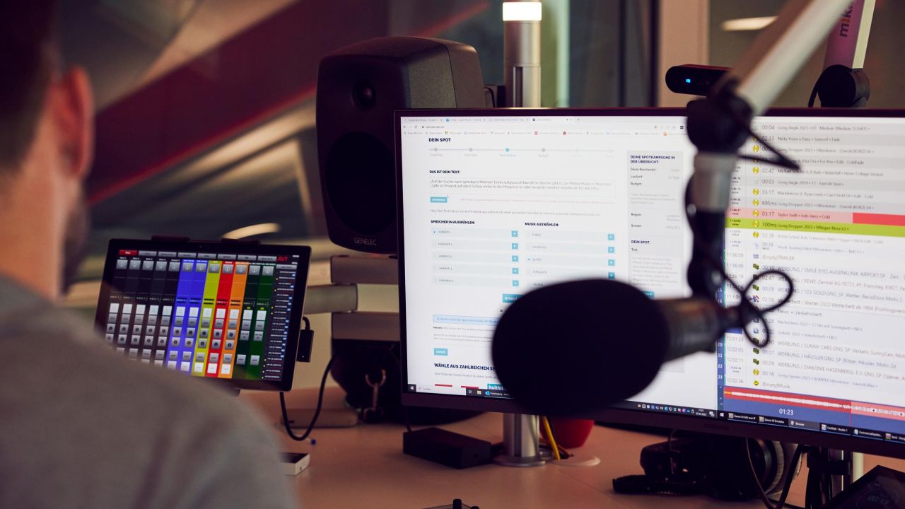 View over the shoulder of a man in a radio studio looking at multiple monitors displaying control interfaces and production timelines